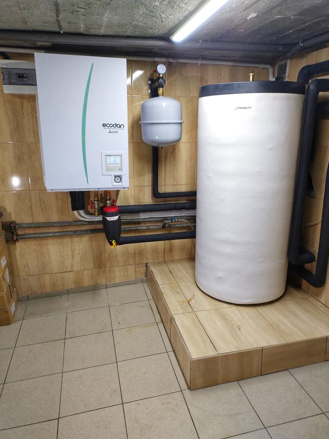Biały zasobnik ciepłej wody użytkowej Trinity i pompa ciepła Ecodan zamontowane na tle beżowych płytek ściennych i podłogowych, widoczne rury instalacyjne.