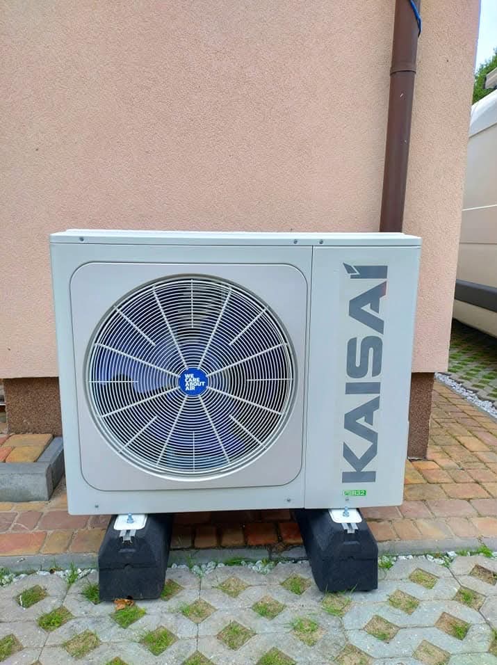 Zewnętrzna jednostka klimatyzacji Kaisai, zamontowana na czarnych gumowych podstawach, ustawiona na betonowej kostce brukowej przed beżową ścianą budynku z rynną spustową.