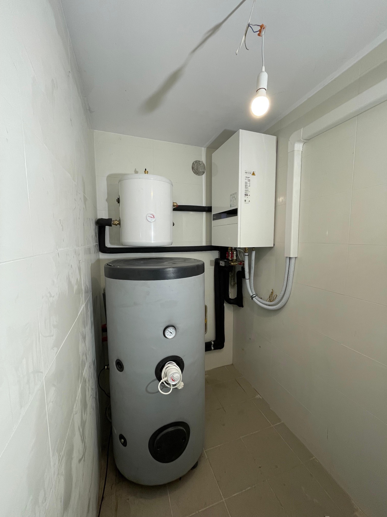 Szary zasobnik ciepłej wody użytkowej z białym zbiornikiem wyrównawczym połączone czarnymi rurami, obok biały piec grzewczy w kotłowni z białymi, karbowanymi przewodami.