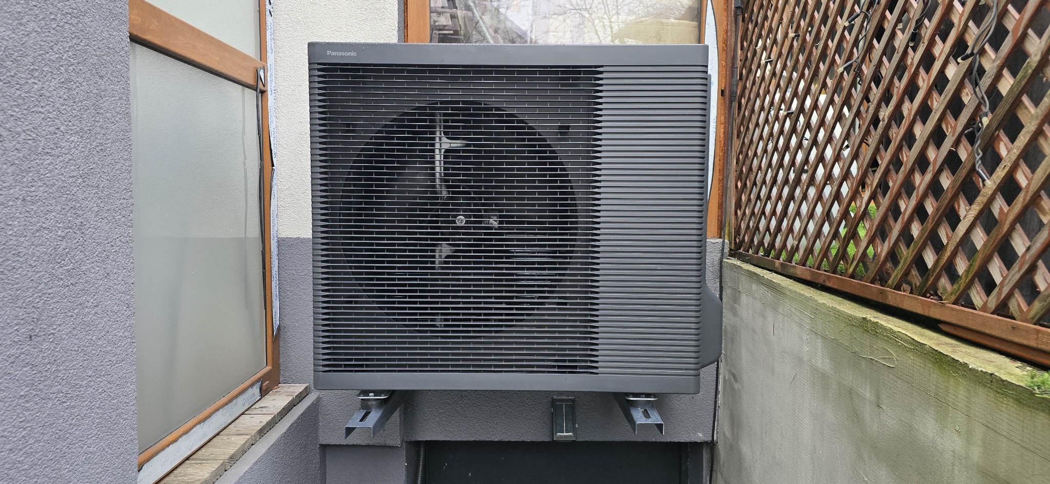 Szara pompa ciepła Panasonic zamontowana na zewnątrz budynku, widoczna kratka wentylacyjna, szara elewacja i drewniana kratownica w tle.