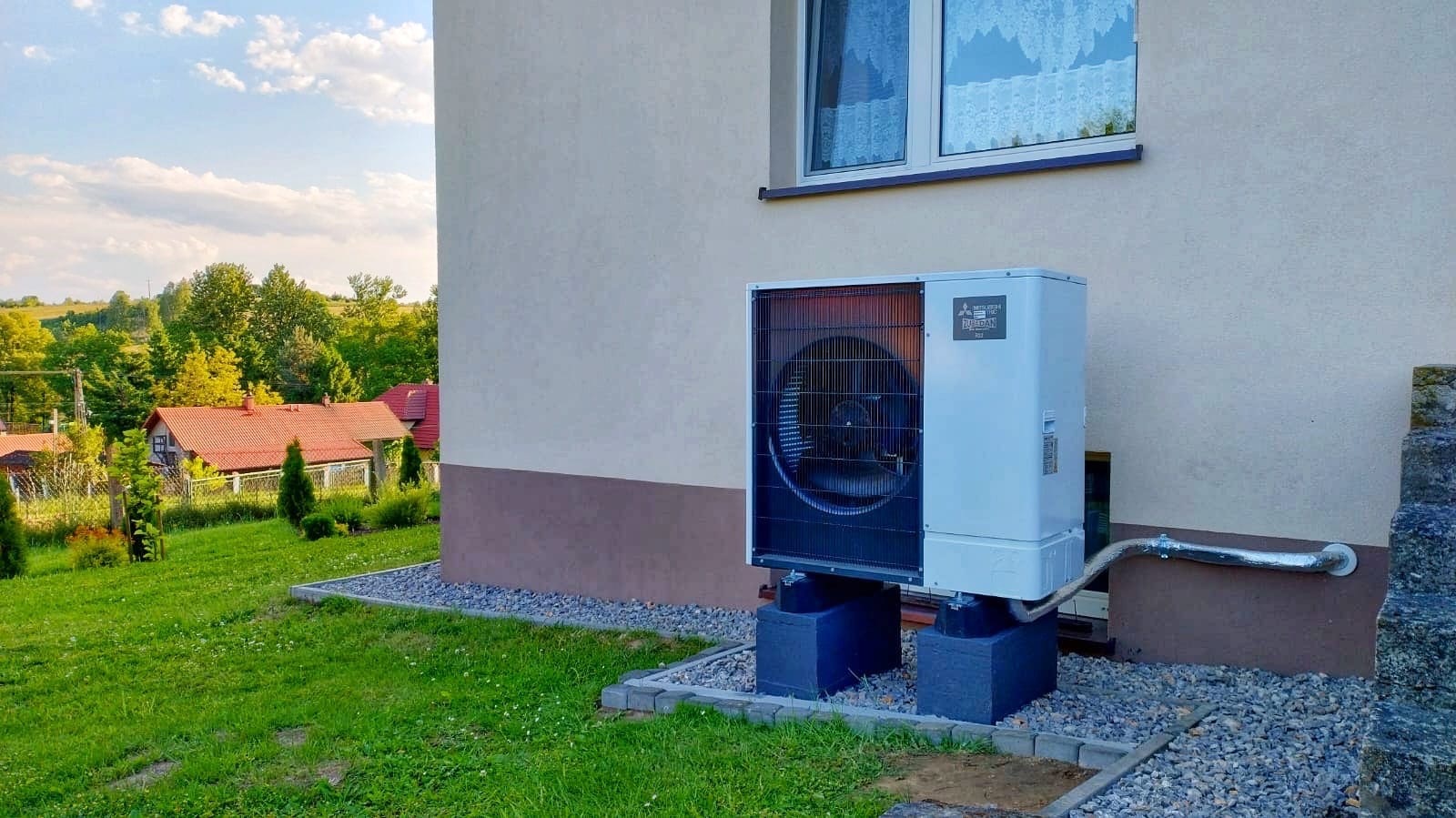 Zewnętrzna jednostka pompy ciepła Mitsubishi Zubadan na betonowych podstawach przy ścianie domu z widokiem na zielony ogród i zabudowania w tle.
