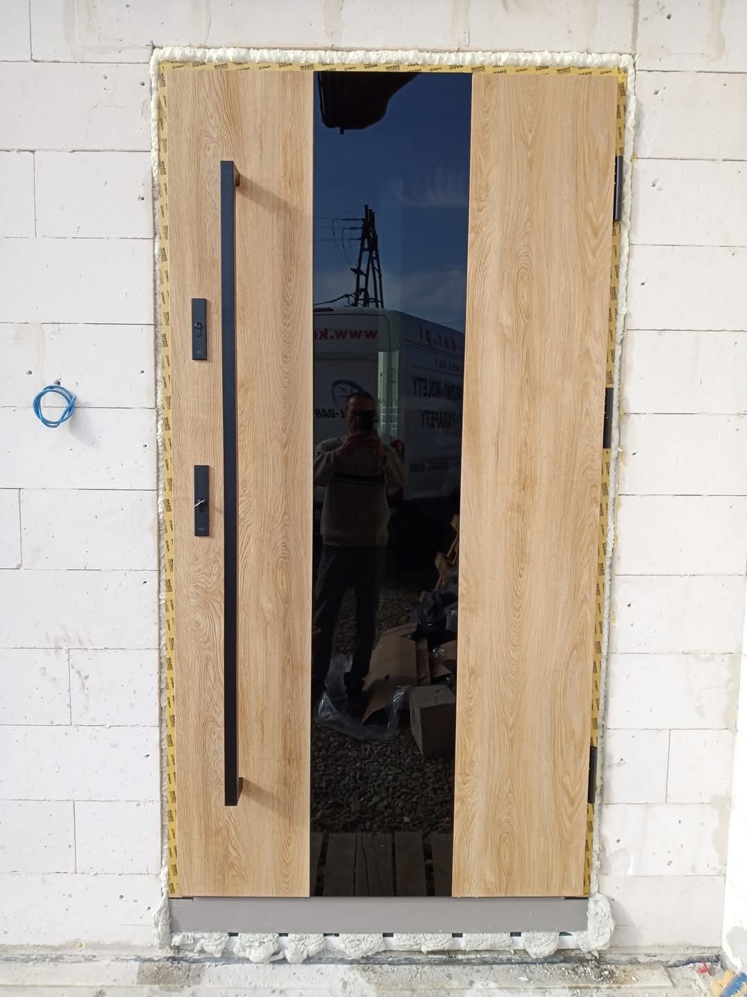 Montaż nowoczesnych drzwi wejściowych z okładziną drewnopodobną i czarnym szkłem, widoczna ościeżnica uszczelniona pianką montażową, ściana z białych bloczków.