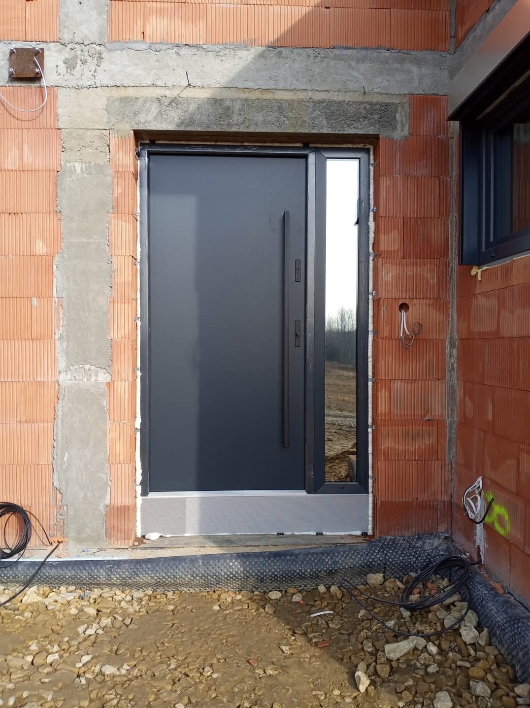 Nowoczesne, antracytowe drzwi wejściowe z bocznym przeszkleniem zamontowane w surowym murze z czerwonej cegły, widoczne przewody elektryczne i fragment okna po prawej stronie.