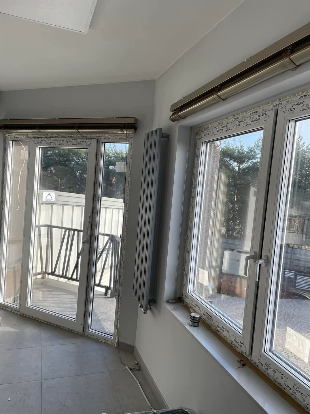 Szary grzejnik panelowy zamontowany na ścianie obok białych okien PCV z widokiem na zewnątrz, z balkonem i zielenią, w jasnym pomieszczeniu z szarą podłogą.