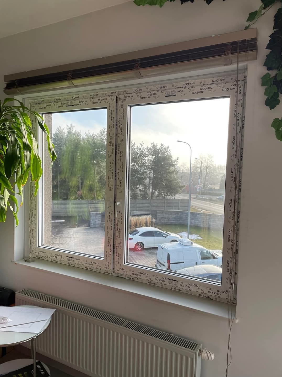 Nowe okno PCV z roletami wewnętrznymi, widok na podjazd z samochodami i drzewa w tle.