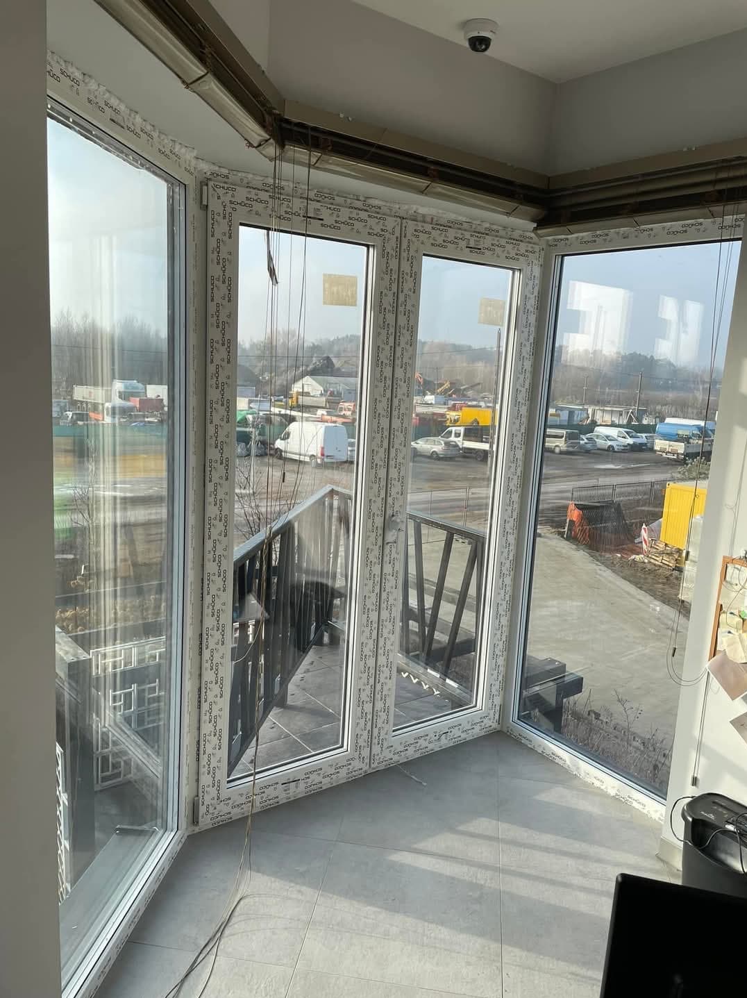 Zabudowany balkon z białymi oknami PCV marki Schuco, widok na plac budowy z kontenerami i maszynami, szare płytki na podłodze, kamera monitoringu na suficie.