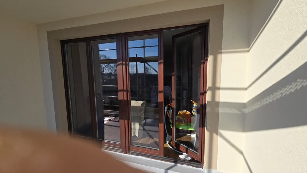 Nowe, brązowe okno balkonowe trzyskrzydłowe, częściowo otwarte, z widokiem na zewnątrz, z jasnymi refleksami słonecznymi na ścianie i podłodze.