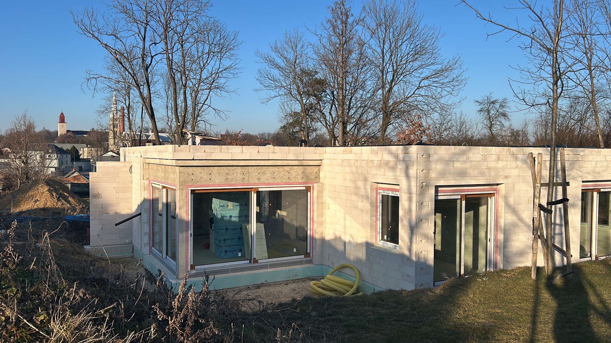 Surowy stan domu z bloczków, widoczne okna z ociepleniem, żółty wąż leży na ziemi, w tle zabudowa miejska z wieżą kościelną.