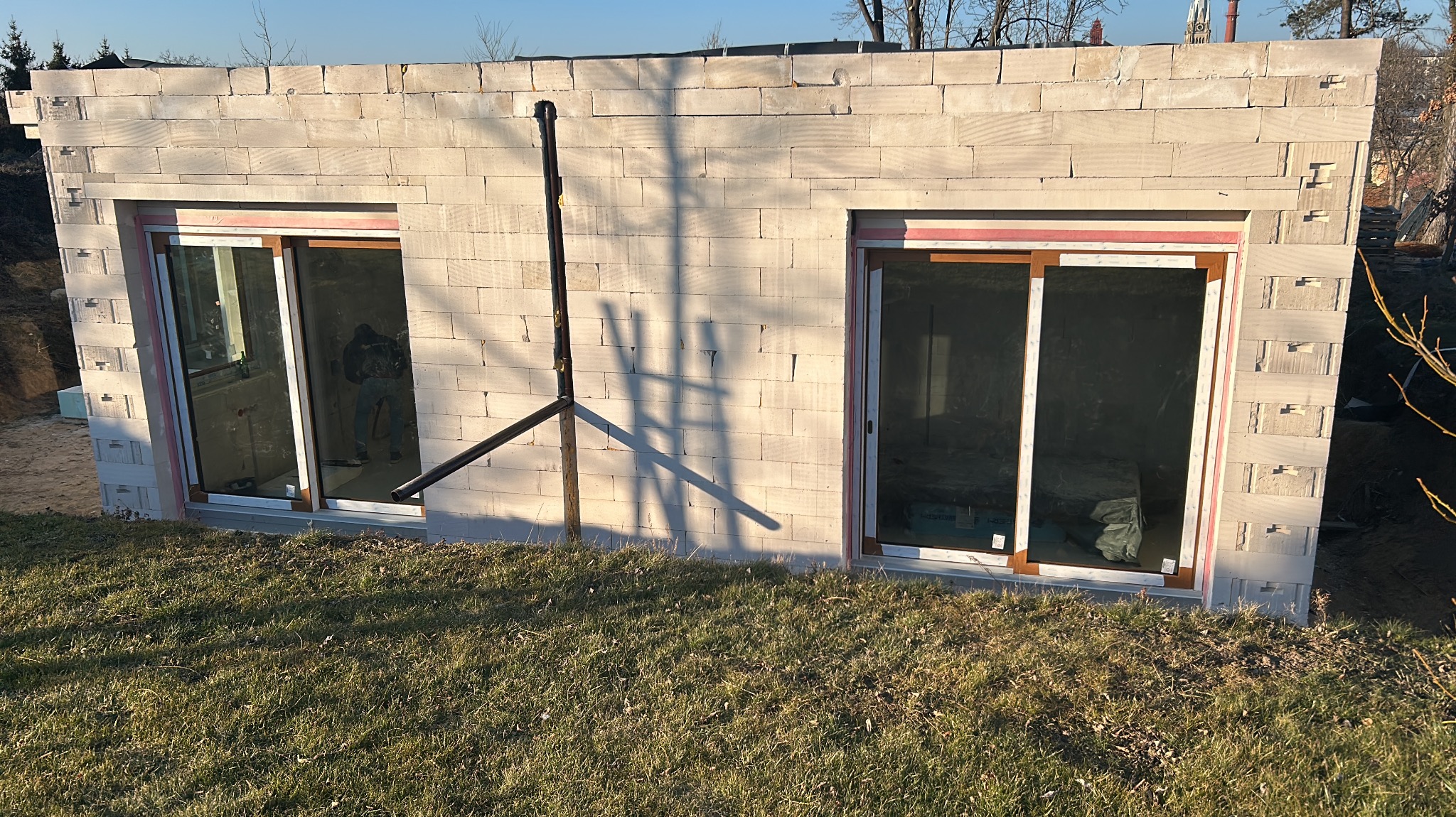 Ściana fundamentowa z bloczków betonu komórkowego, z dwoma oknami z brązowymi ramami, widoczne ocieplenie pionowe po bokach okien, cień drabiny na ścianie, trawa na pierwszym planie.