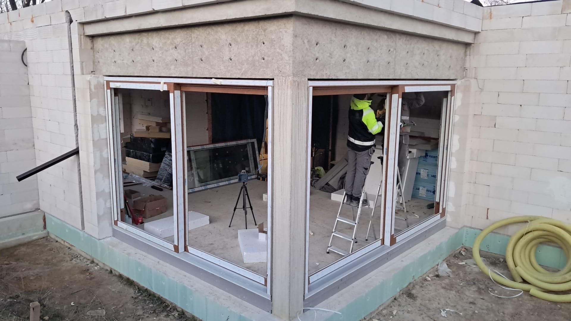 Montaż narożnego okna panoramicznego w surowym stanie, z widocznym pracownikiem na drabinie, laserem na statywie i materiałami budowlanymi wewnątrz.