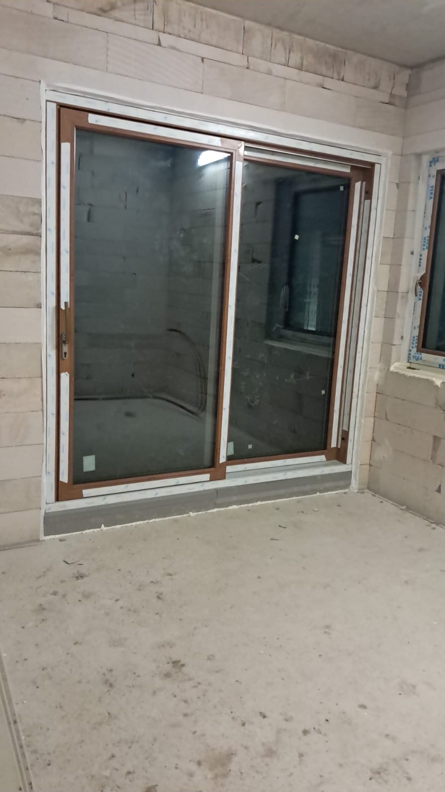 Zamknięte, przesuwne okno balkonowe z brązową ramą, zamontowane w surowym wnętrzu z białych bloczków, zabezpieczone taśmą malarską.