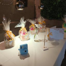 Restauracja Veneto - Dekoracje stołu na przyjęciu: koszyczki z upominkami, kwiaty w wazonie, księga gości i błękitny aparat Instax, oświetlone wiszącymi lampami.