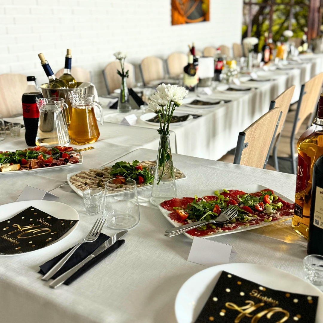 Uroczysty stół z białym obrusem, zastawiony talerzami, sztućcami, napojami, dekoracjami kwiatowymi i przekąskami na przyjęcie urodzinowe.