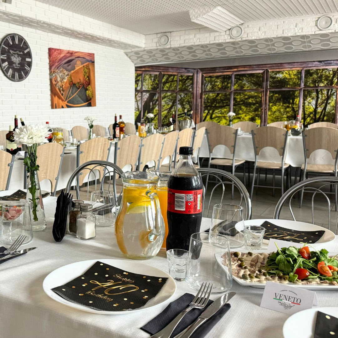 Uroczysty stół z dekoracjami urodzinowymi, nakryty białym obrusem, z napojami, sałatką i eleganckimi serwetkami. W tle widok na salę z oknami.