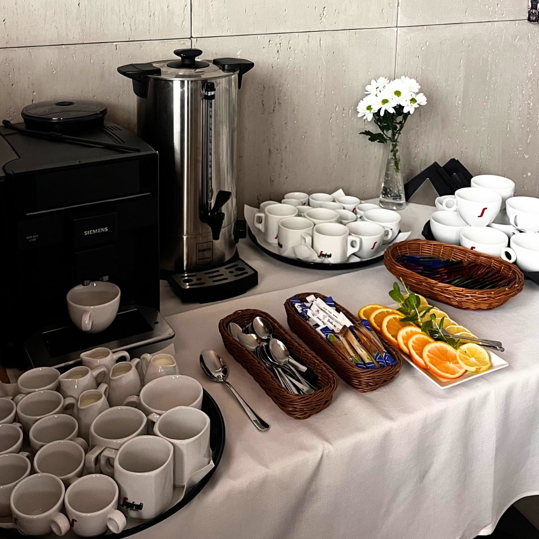 Stół z cateringiem kawowym: ekspres Siemens, termos, filiżanki, owoce i dodatki. Elegancki bufet kawowy z białym obrusem i kwiatami.