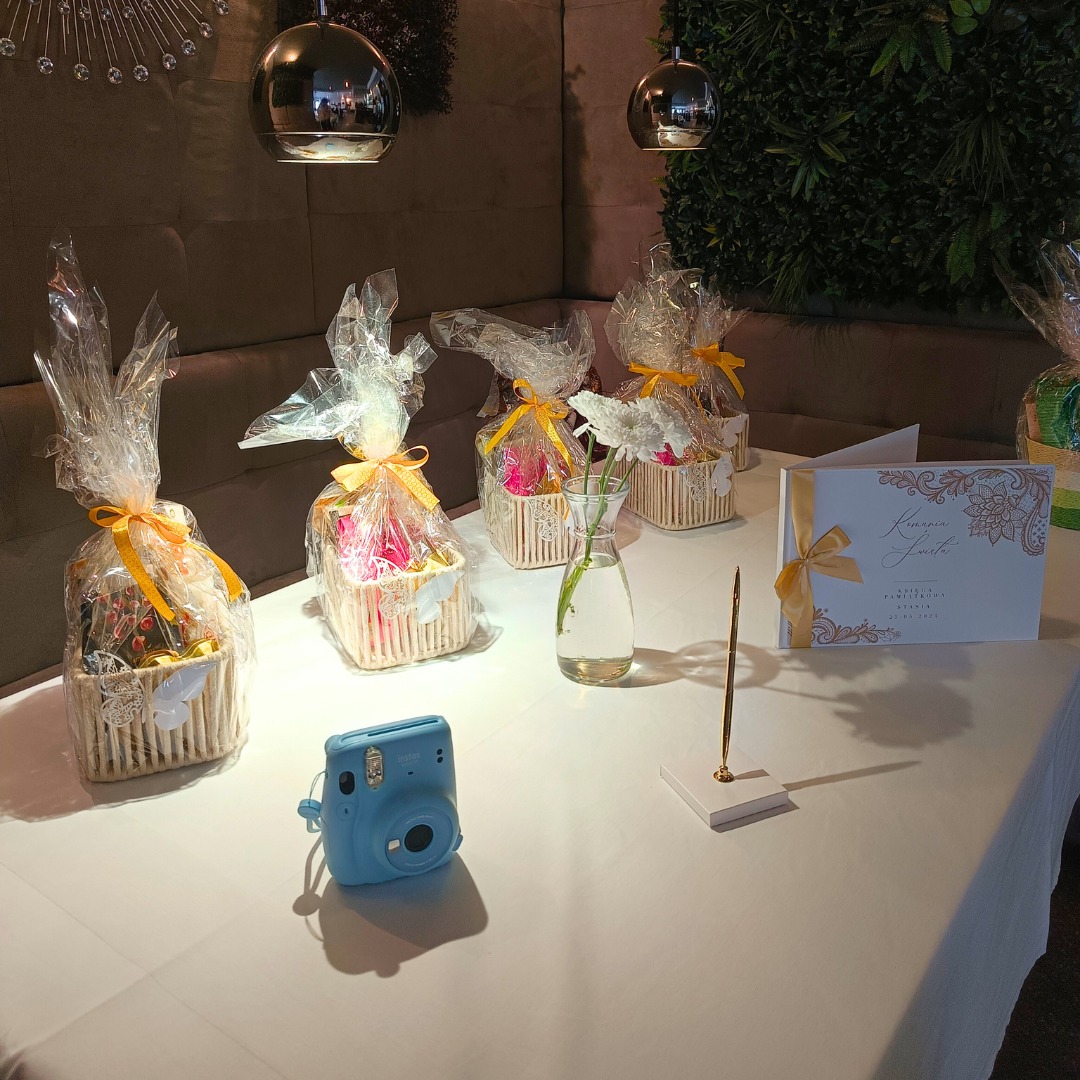 Dekoracje stołu na przyjęciu: koszyczki z upominkami, kwiaty w wazonie, księga gości i błękitny aparat Instax, oświetlone wiszącymi lampami.