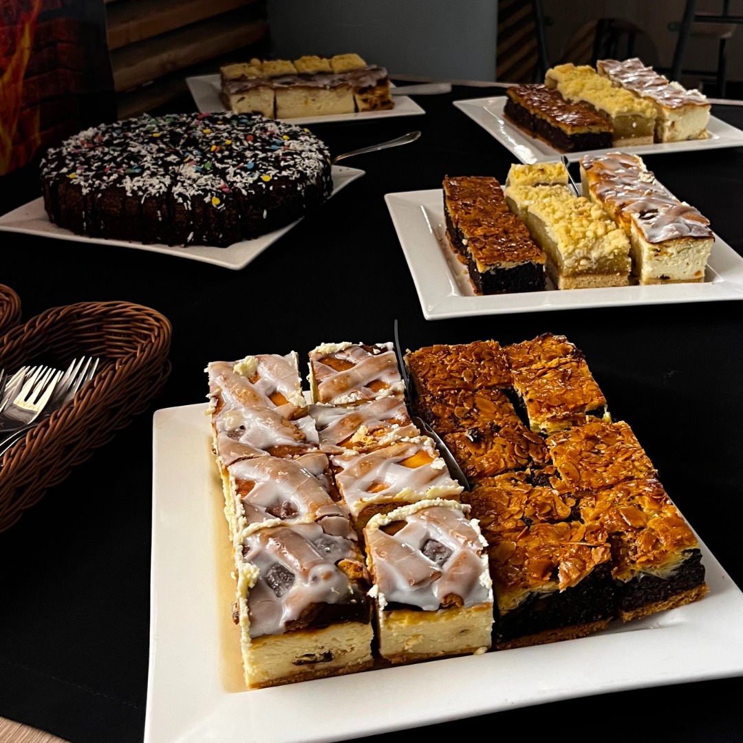 Urozmaicony wybór ciast na białych talerzach, w tym mazurek, sernik i ciasto czekoladowe z posypką, ustawionych na czarnym stole. Widoczny koszyk z widelcami.