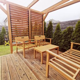 ProCraft - Zadaszony taras z drewnianą podłogą i meblami ogrodowymi z jasnego drewna, w tle rząd zielonych tuj i drewniany płot, z boku widoczna myjka ciśnieniowa.