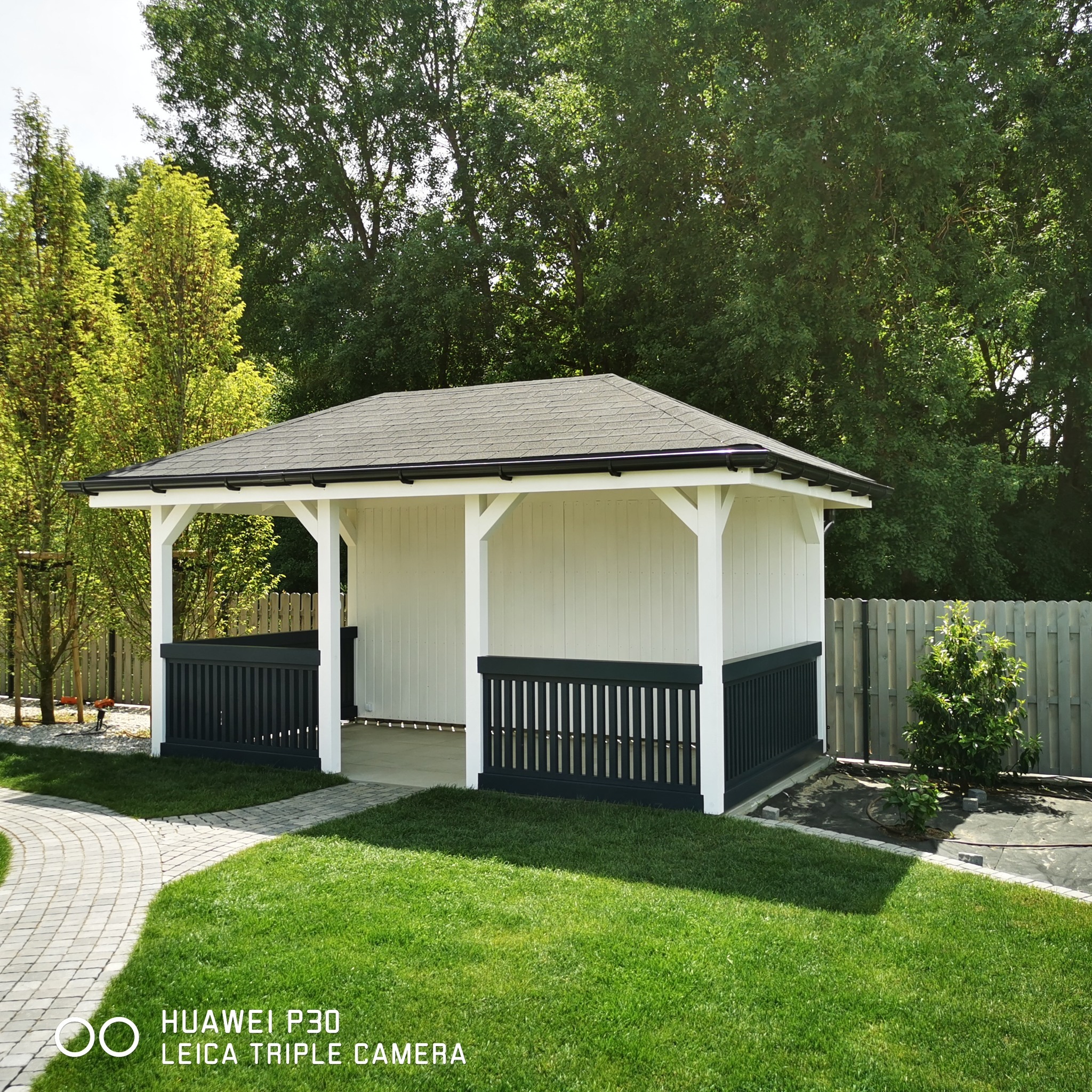 Biała altana z ciemnoszarymi balustradami i dachem z gontu bitumicznego, otoczona zielonym trawnikiem i drzewami.