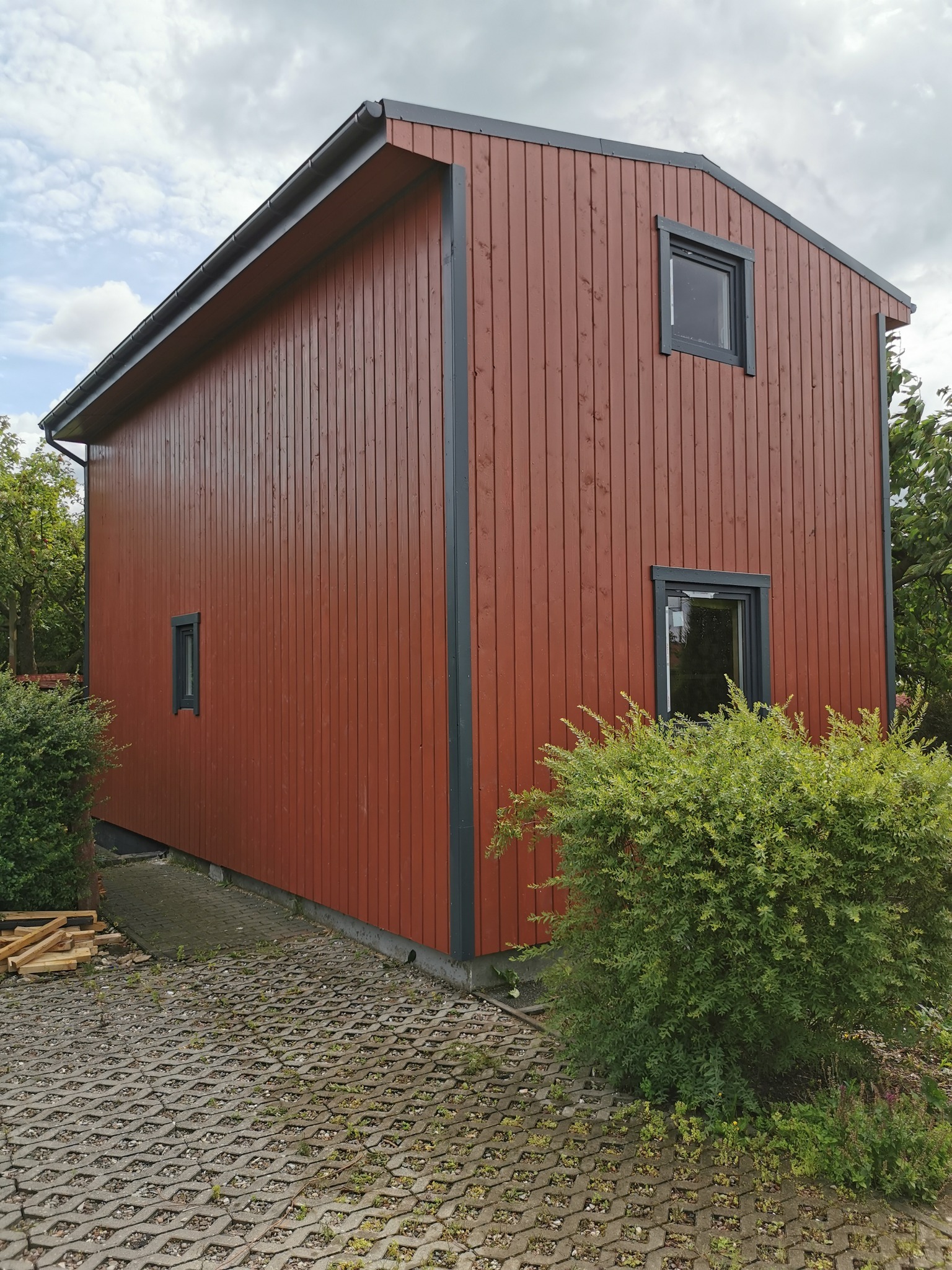 Dwukondygnacyjny budynek z drewnianą elewacją w kolorze czerwono-brązowym i szarymi obramowaniami okien, otoczony zielenią i wyłożony ażurową kostką brukową.