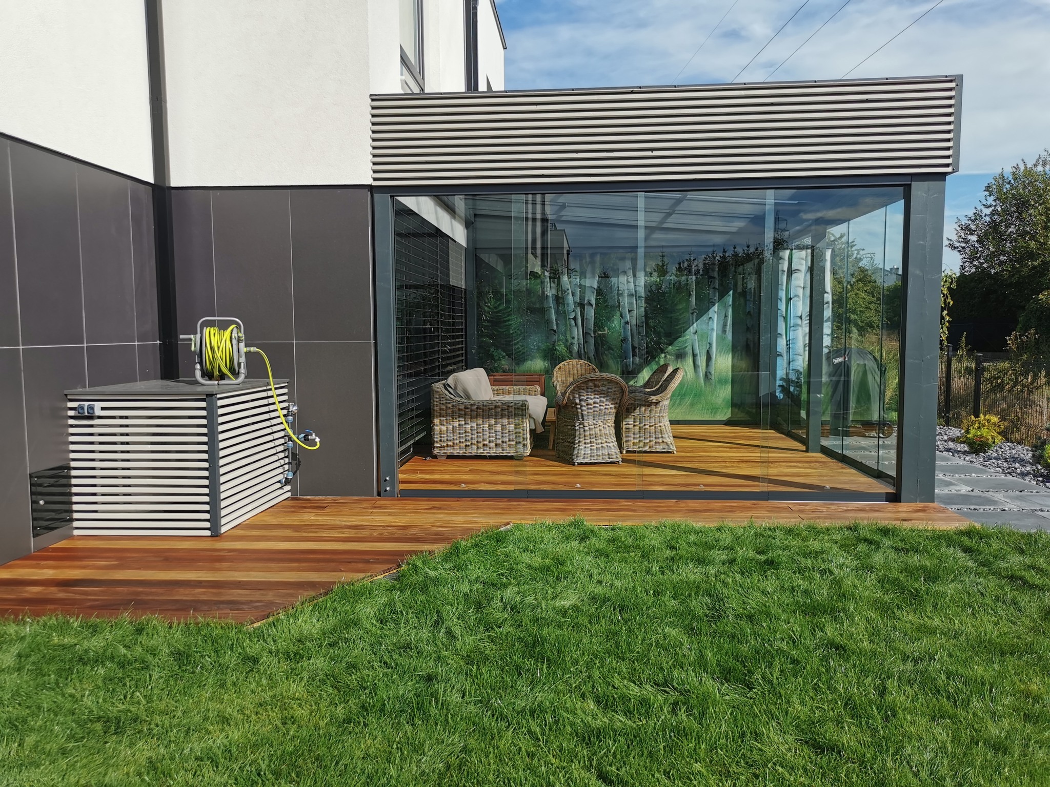Zadaszony taras z drewnianą podłogą i szklanymi ścianami, wyposażony w rattanowe meble. Na zewnątrz widoczny trawnik, nowoczesna elewacja budynku z szarą okładziną i zwinięty wąż ogrodowy na boku.