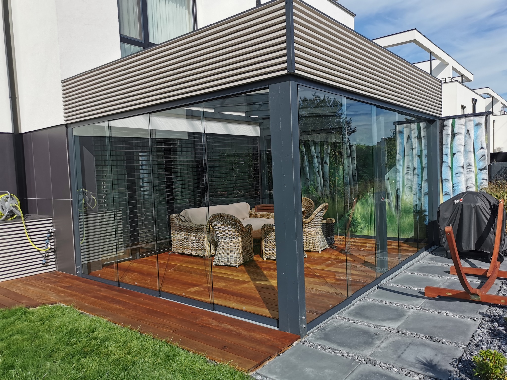 Nowoczesna przeszklona konstrukcja z drewnianą podłogą i meblami rattanowymi, z widokiem na ogród i elementami dekoracyjnymi na ścianie.