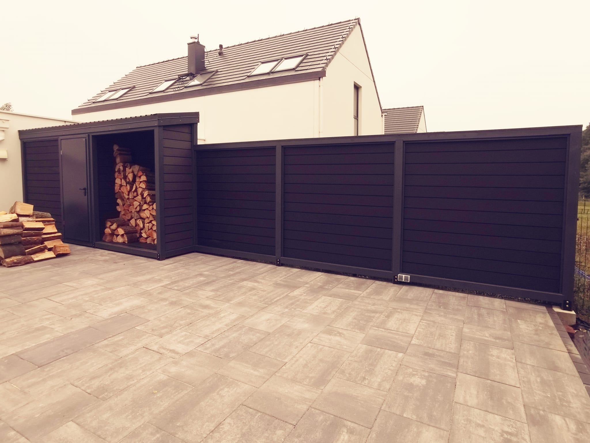Nowoczesna, czarna konstrukcja z poziomych desek, pełniąca funkcję osłony i składu drewna, na tle domu z szarym dachem i beżową elewacją, ułożona na jasnej kostce brukowej.