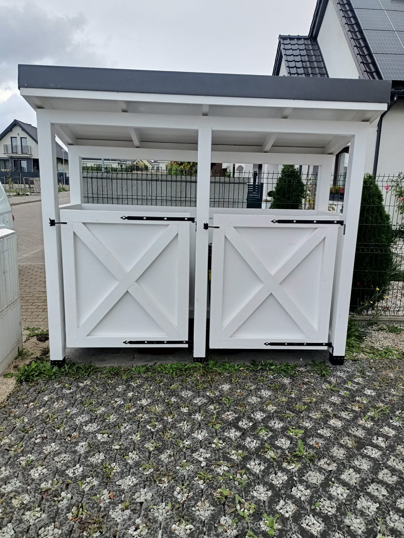 Biała drewniana konstrukcja z dwoma zamykanymi drzwiczkami z krzyżowym wzorem, prawdopodobnie osłona na pojemniki na śmieci, ustawiona na ażurowej kostce brukowej z wyrastającą trawą, w tle dom...