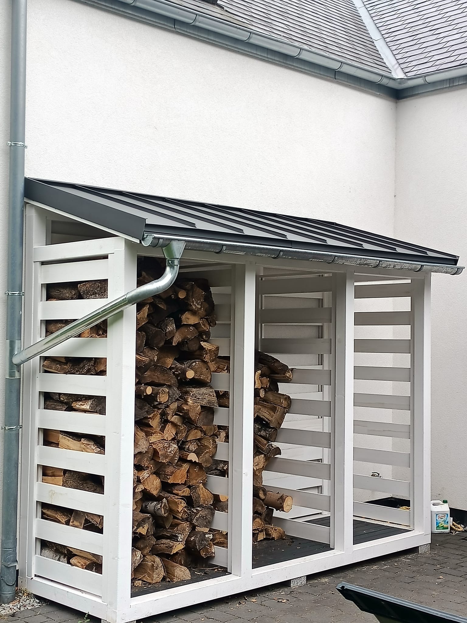 Drewniana altana na drewno opałowe z szarym dachem blaszanym i rynną, wypełniona pociętymi kawałkami drewna, stojąca przy białej ścianie domu z widoczną rurą spustową.
