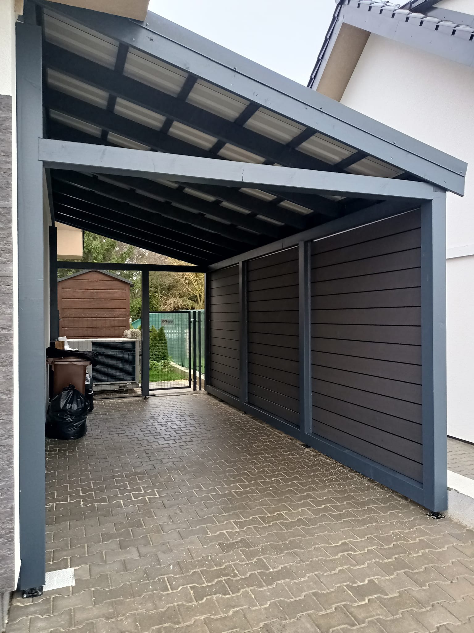 Zadaszona konstrukcja z szarymi elementami i brązowymi panelami bocznymi, widziana z zewnątrz na tle kostki brukowej i fragmentu budynku.