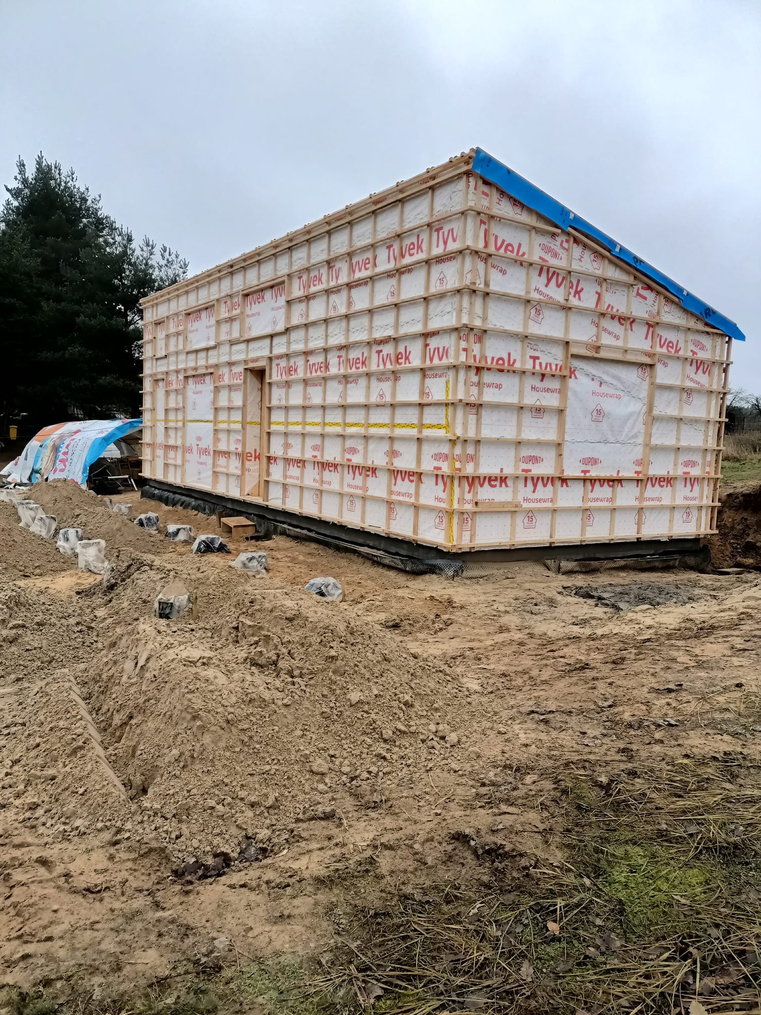 Budowa domu szkieletowego w trakcie wznoszenia, widoczne drewniane ramy ścian pokryte membraną Tyvek, przygotowane fundamenty i wykop pod instalacje, pochmurne niebo.