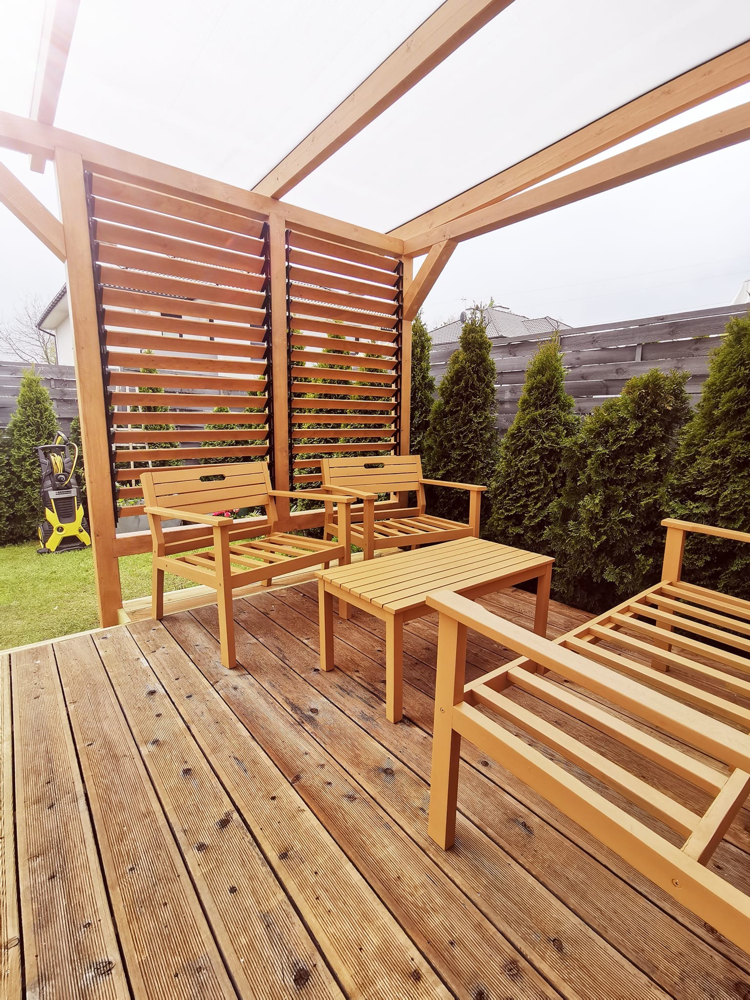 Zadaszony taras z drewnianą podłogą i meblami ogrodowymi z jasnego drewna, w tle rząd zielonych tuj i drewniany płot, z boku widoczna myjka ciśnieniowa.