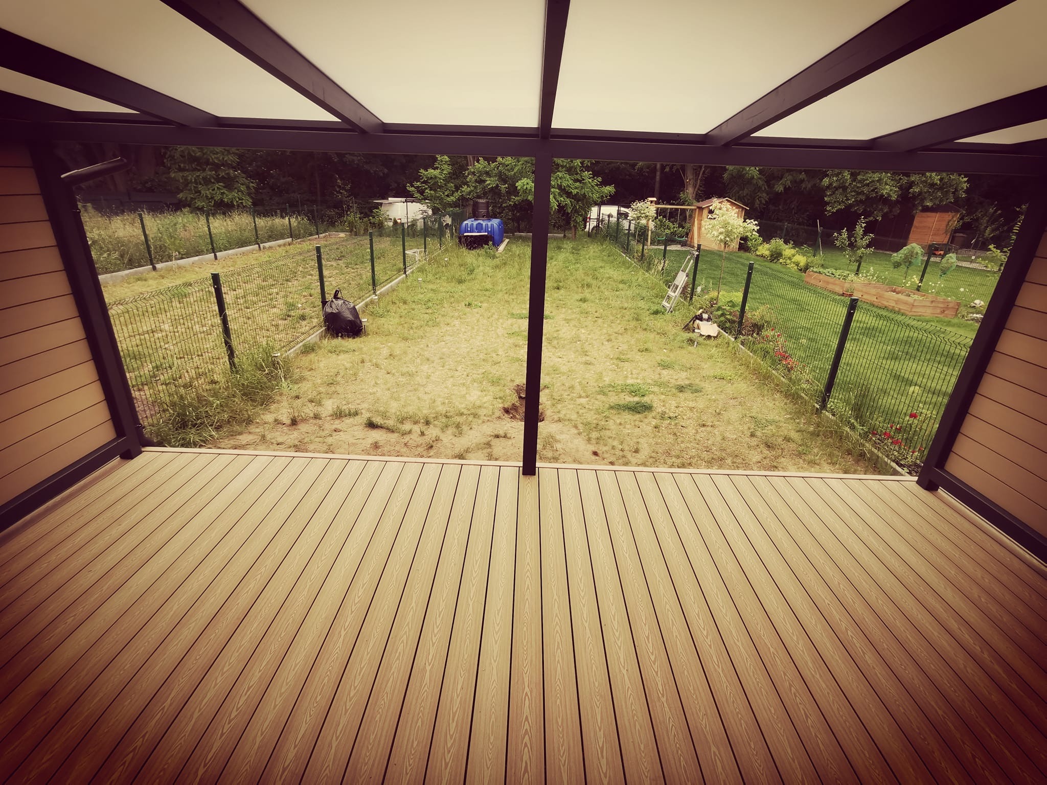 Zadaszony taras z desek kompozytowych, widok na zarośnięty ogród z siatkowym ogrodzeniem i elementami wyposażenia, perspektywa z wnętrza konstrukcji.