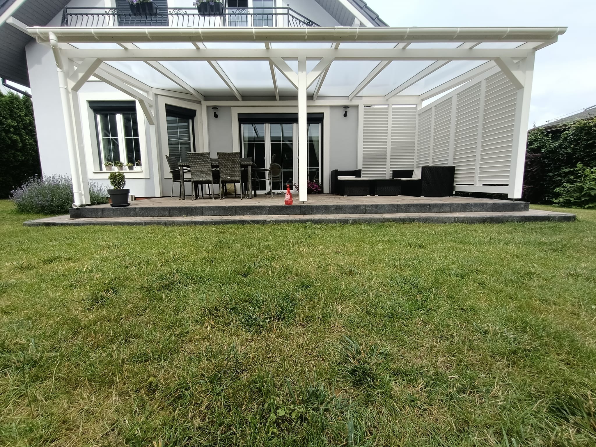 Nowoczesna, biała konstrukcja z zadaszeniem, tworząca zadaszony taras w miejscowości Dobra, z widocznym zestawem mebli ogrodowych i częściowo osłoniętą ścianką z poziomych listew, umieszczona...