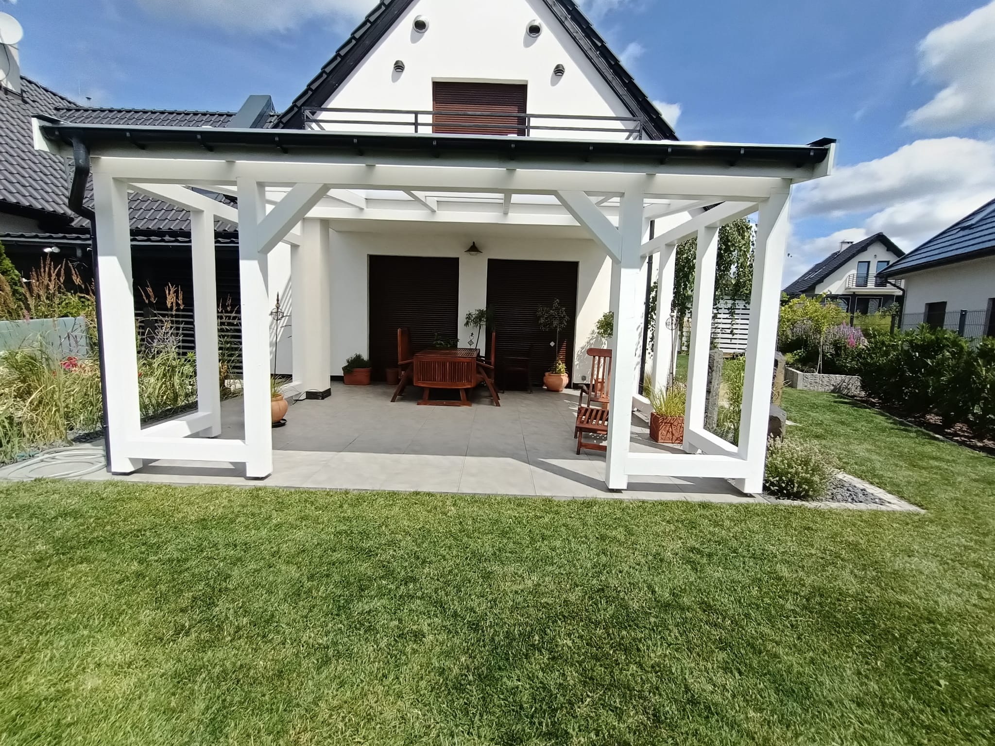 Biała pergola z płaskim dachem przylegająca do białego domu z ciemnymi okiennicami, widok z ogrodu na drewniany stół i krzesła wewnątrz.