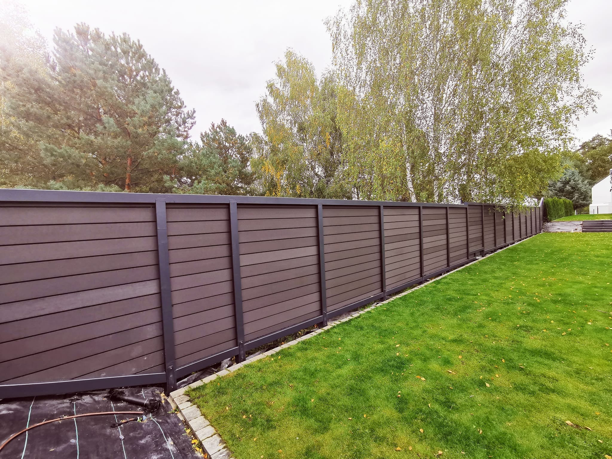 Długi, nowoczesny płot kompozytowy w kolorze ciemnego drewna, z metalowymi słupkami, oddzielający trawnik od drzew w tle, widoczne elementy instalacji nawadniającej.