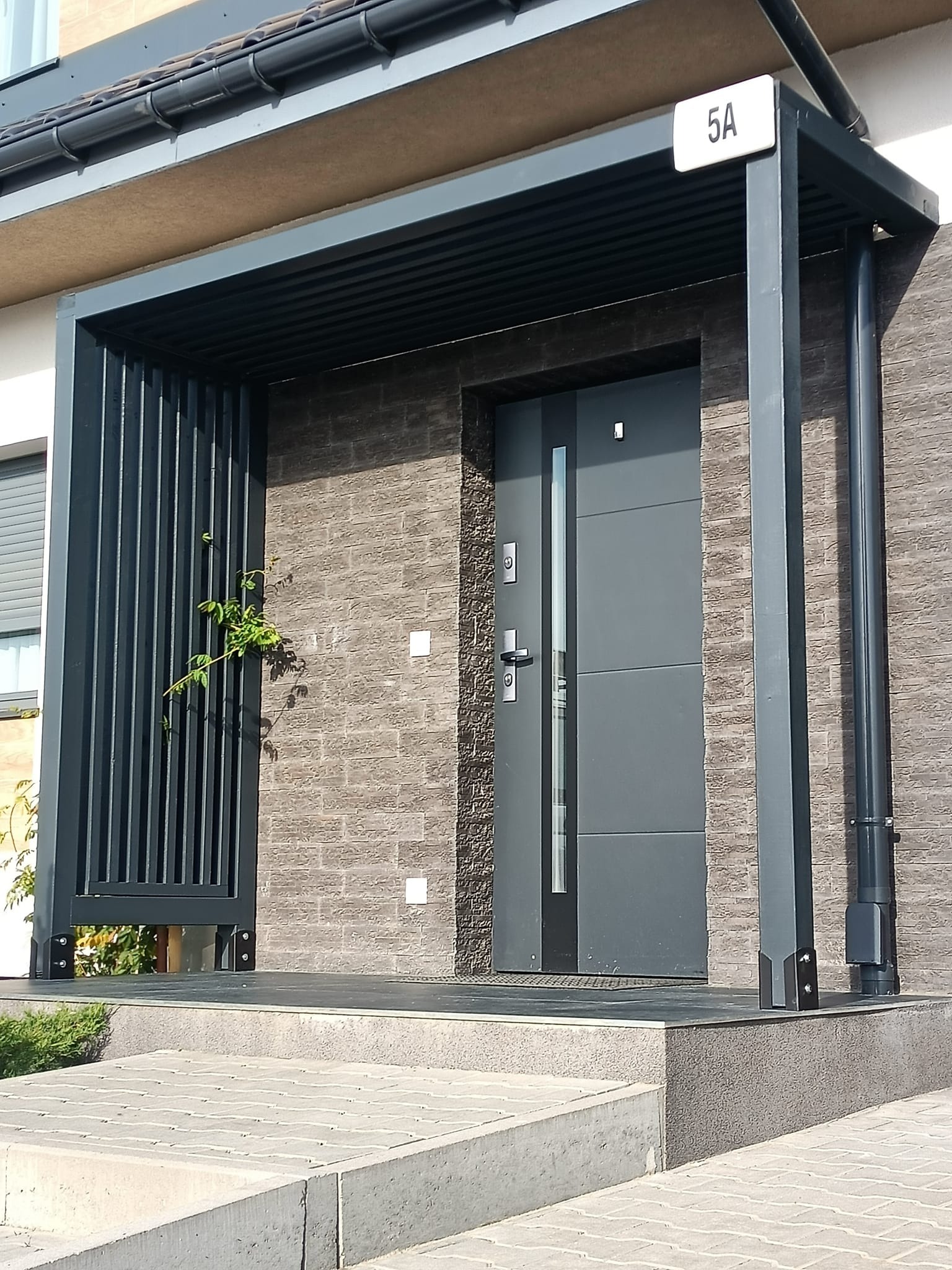 Nowoczesne zadaszenie nad wejściem do domu z szarej konstrukcji stalowej, z numerem domu 5A, z elementami pionowych listew i pnącą się rośliną.
