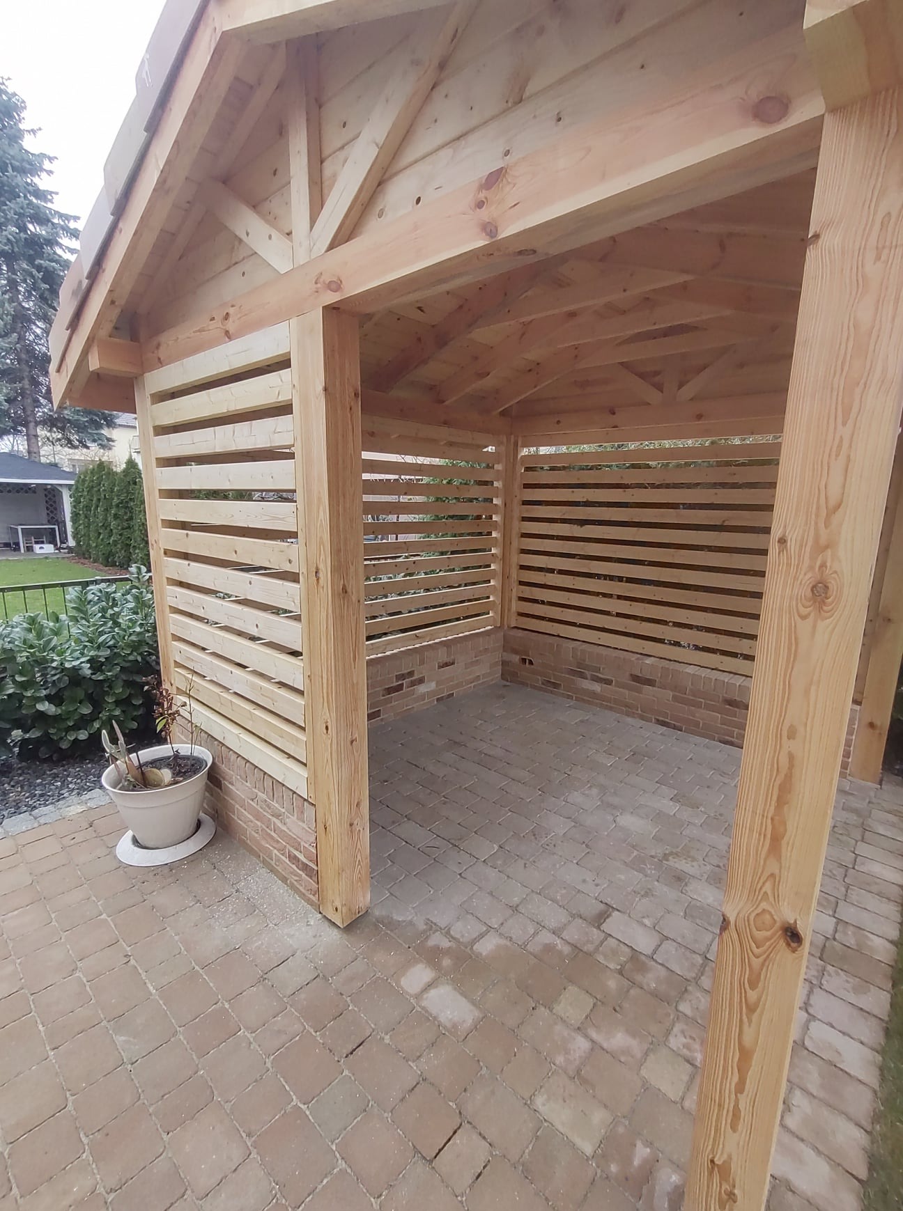 Świeżo wybudowana drewniana konstrukcja z poziomymi listwami i ceglaną podstawą, widok z zewnątrz na kamienną podłogę, donica z rośliną przy jednej ze ścian.