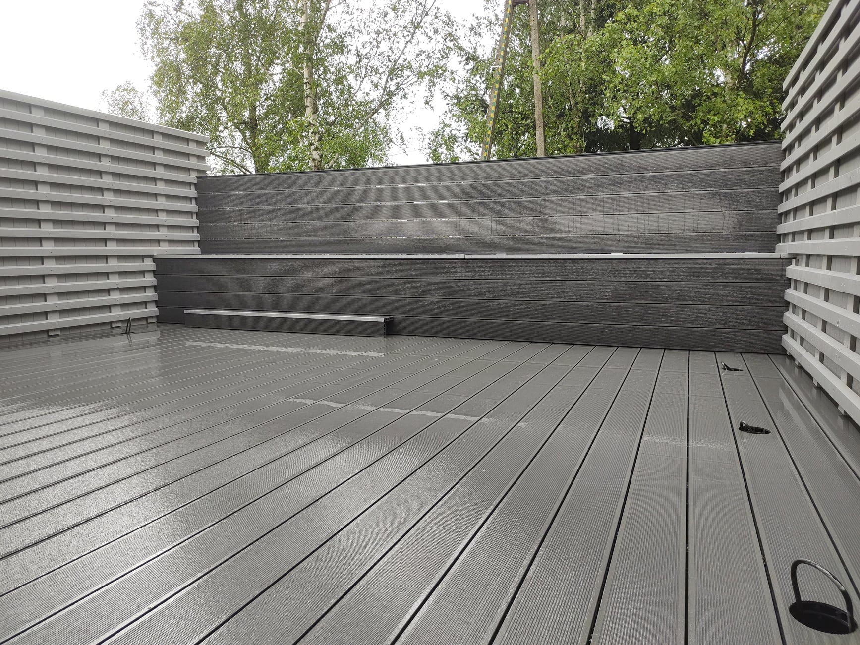 Nowoczesny taras z szarych desek kompozytowych z niskim, ciemnoszarym siedziskiem i jasnoszarym ażurowym panelem bocznym, widok z góry na mokrą powierzchnię po deszczu.