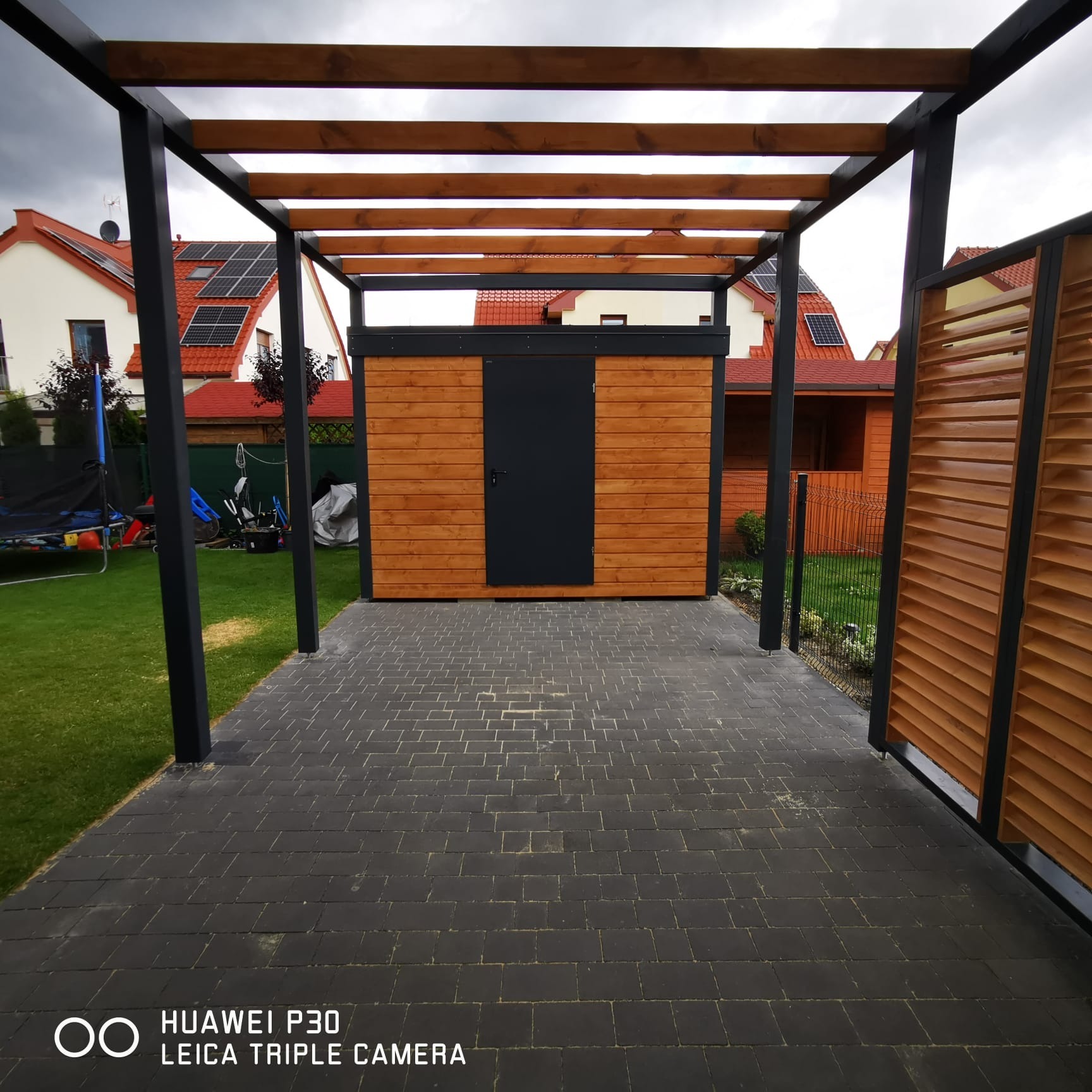 Nowoczesna pergola z czarnymi metalowymi słupami i drewnianym zadaszeniem, obok drewniany schowek z czarnymi drzwiami, na tle kostki brukowej i zielonego trawnika.