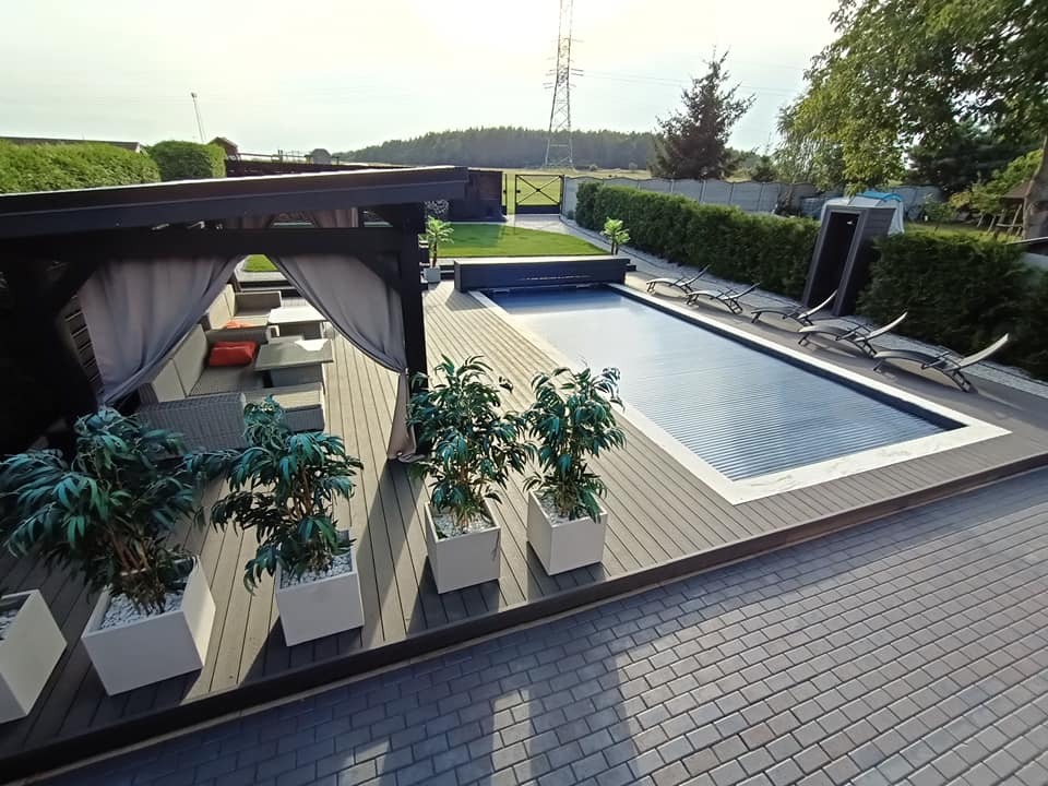 Zadaszony taras z szarej deski kompozytowej, zadaszenie z zasłonami, basen z roletą, leżaki i zadbany ogród w tle.
