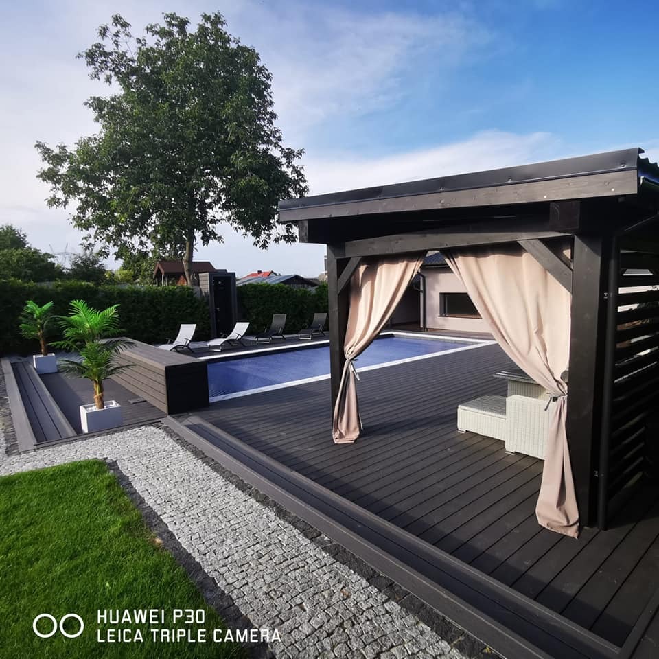 Nowoczesny taras kompozytowy z ciemnego drewna, przylegający do basenu z niebieską pokrywą, z widoczną altaną z zasłonami, ścieżką z jasnych kamieni i dekoracyjnymi palmami w donicach.