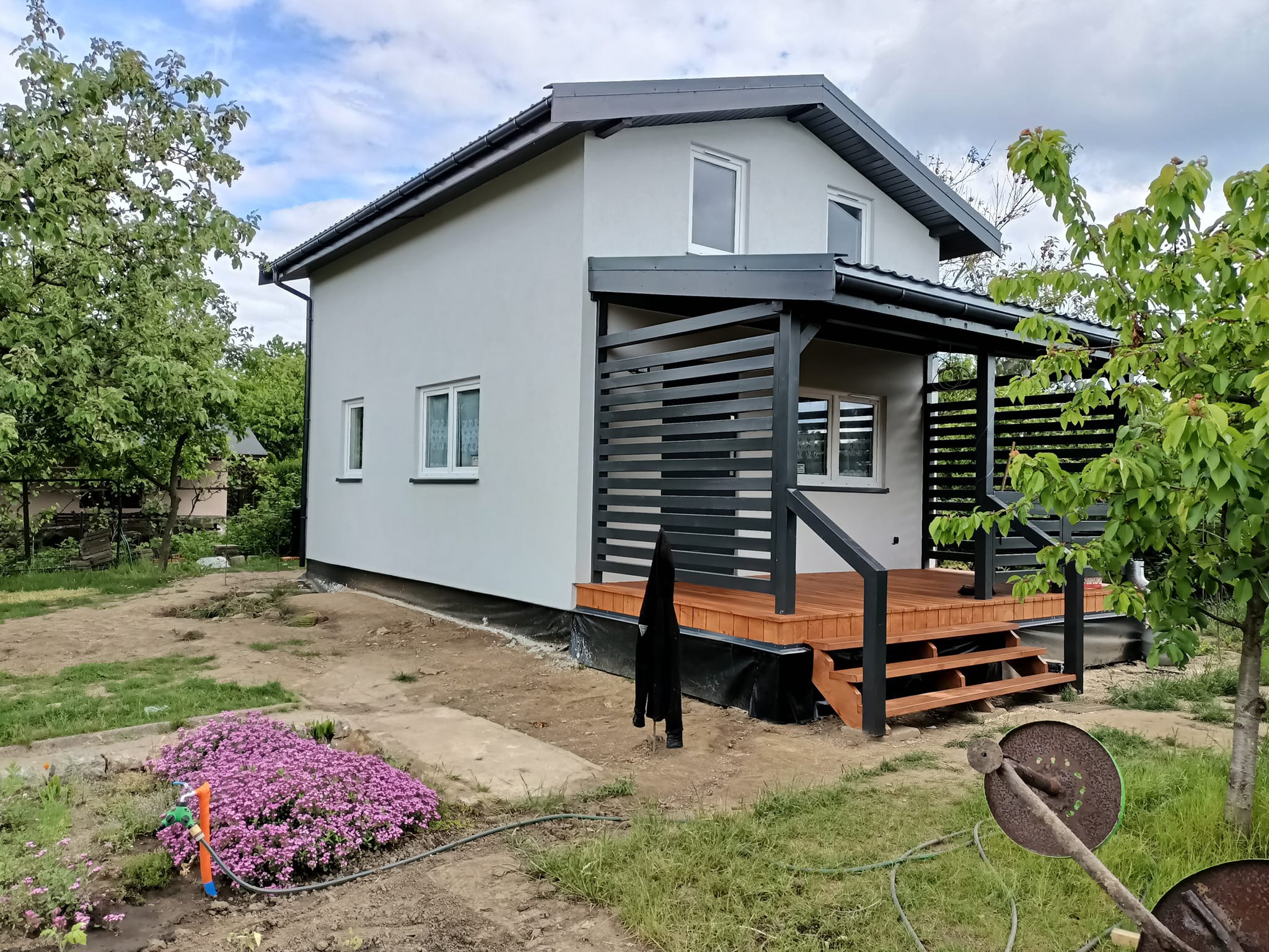 Nowoczesny, szary dom z drewnianym tarasem i czarną pergolą, widok z zewnątrz, ogród w trakcie aranżacji, narzędzia ogrodnicze leżą na trawie.