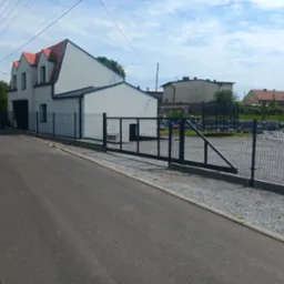 Nowoczesna posesja z grafitowym ogrodzeniem panelowym i przesuwną bramą. Widok na dom z czerwonym dachem i grafitową bramą wjazdową. Całość dopełnia szara nawierzchnia.