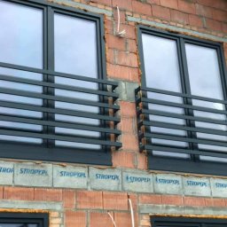 Richly-Spaw - Dwa okna z czarnymi ramami i metalowymi balustradami ochronnymi na surowej ceglanej ścianie budynku w trakcie budowy. Widoczne detale montażu.