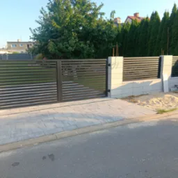 Nowoczesna, pozioma brama wjazdowa z metalowymi lamelami w kolorze grafitowym, osadzona na jasnych, betonowych słupkach. Widoczne kostki brukowe i zieleń w tle.