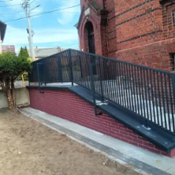 Rampa dla niepełnosprawnych z czarną, metalową balustradą przy ceglanym budynku. Widoczny fragment chodnika i rabata z drzewem. Słoneczny dzień, błękitne niebo.