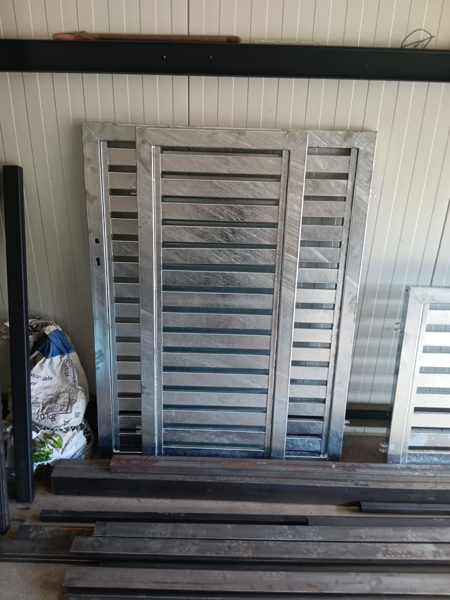 Surowa, metalowa brama z poziomymi szczeblami, w trakcie produkcji lub składowania, z elementami konstrukcyjnymi i workiem obok.