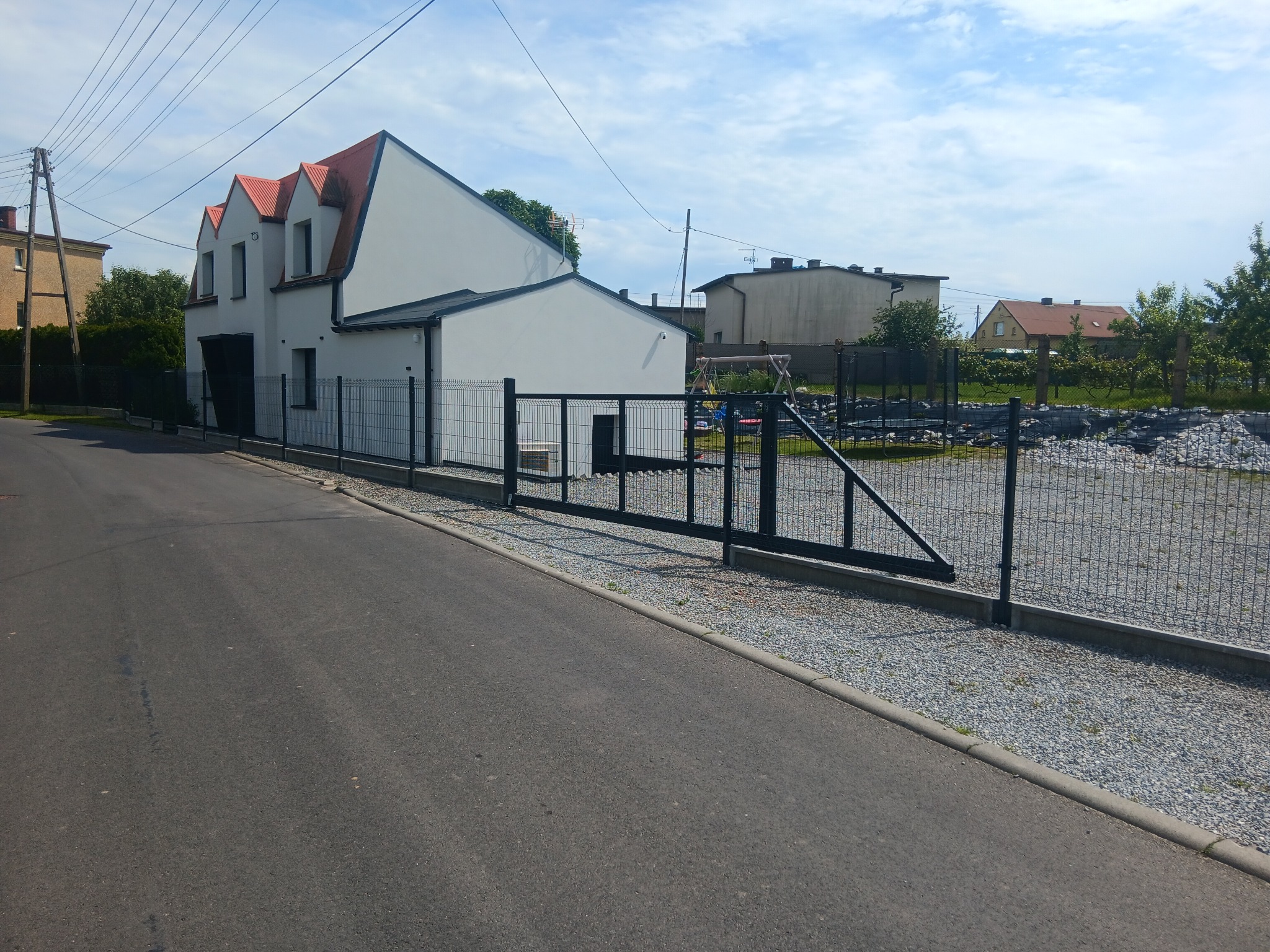 Nowoczesna posesja z grafitowym ogrodzeniem panelowym i przesuwną bramą. Widok na dom z czerwonym dachem i grafitową bramą wjazdową. Całość dopełnia szara nawierzchnia.