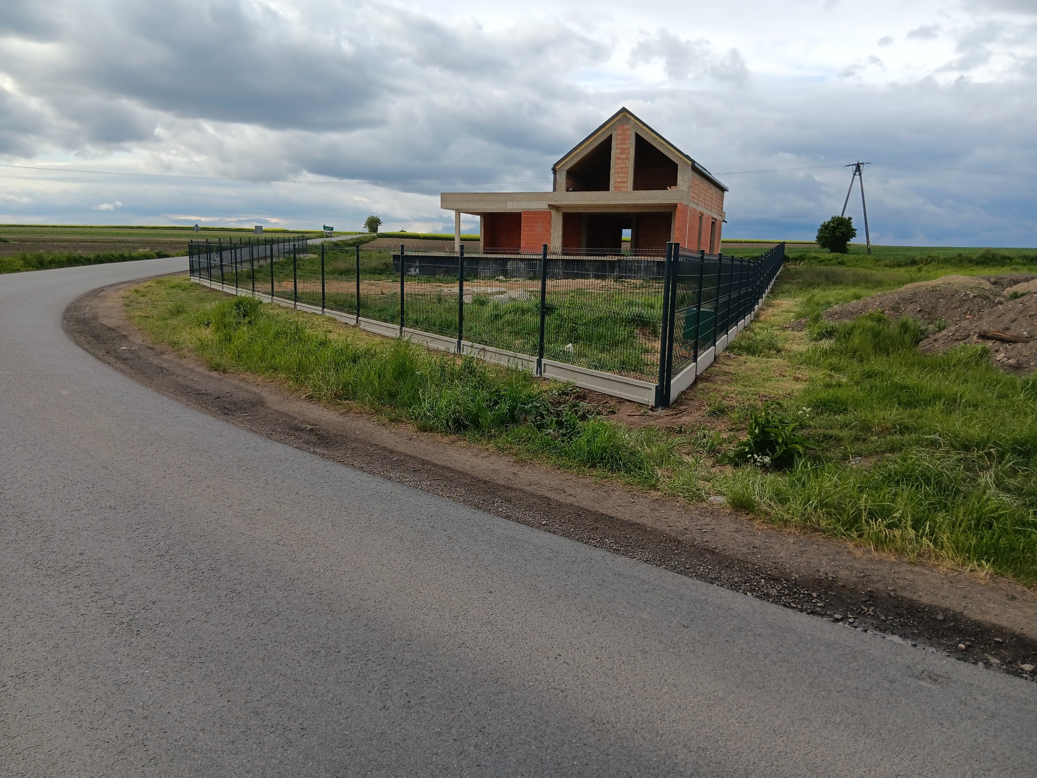 Nowoczesne ogrodzenie panelowe okala plac budowy domu jednorodzinnego na tle pochmurnego nieba i zielonych pól. Widoczne słupki i podmurówka.